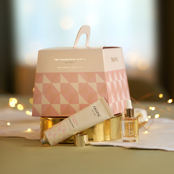 Tropic Frangipani Santal hand cream and restoring face oil beside the pink geometric seasonal gift bag with fairy lights.