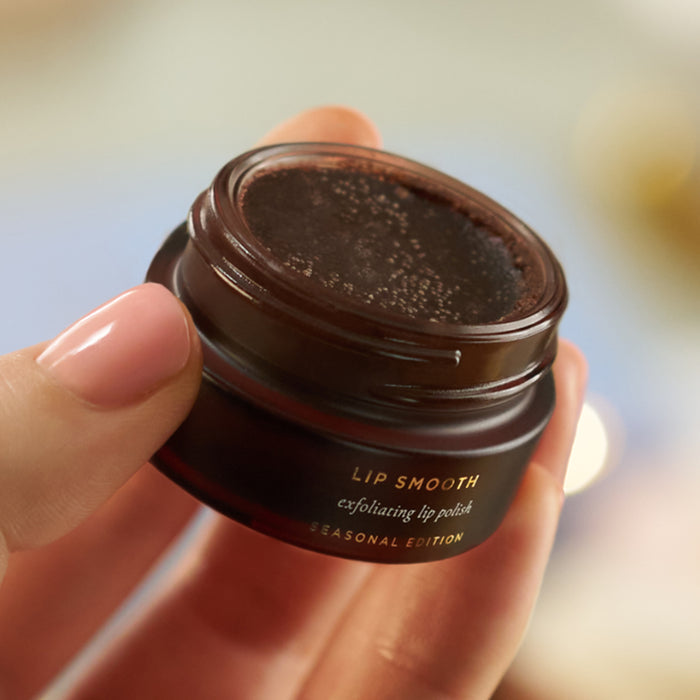 Hands holding open Tropic Lip Smooth exfoliating lip polish jar showing the dark sugar scrub texture.