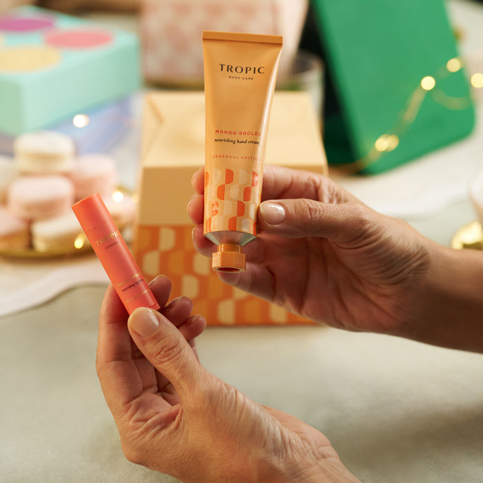 Hands holding Tropic Mango Brûlée nourishing hand cream and lip balm against a festive gift backdrop.