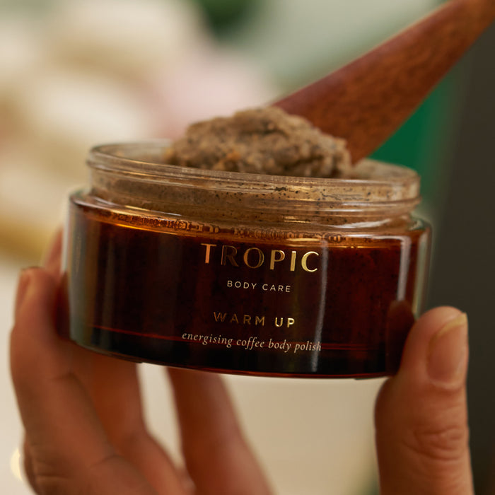 Hands holding open jar of Tropic Warm Up Energising Coffee Body Polish with wooden spatula.